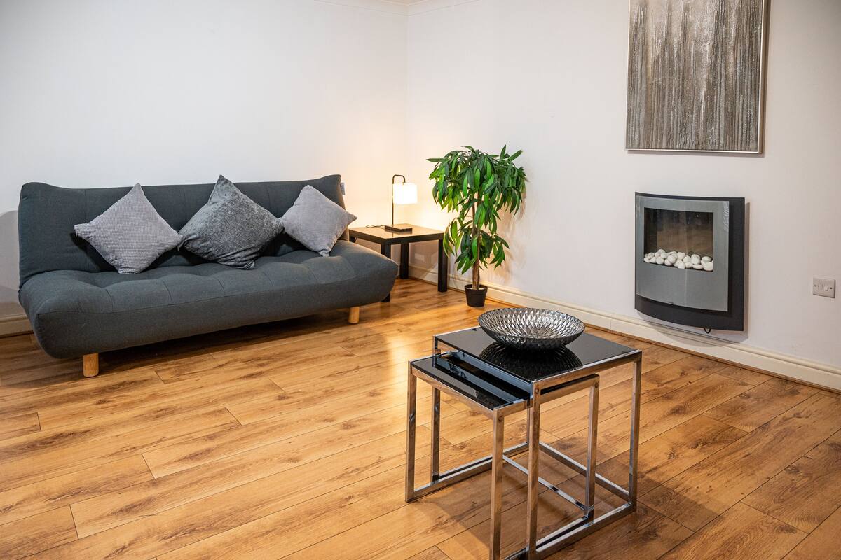 A modern living room with a grey sofa, a small side table, a floor lamp, a potted plant, and a television on the wall.