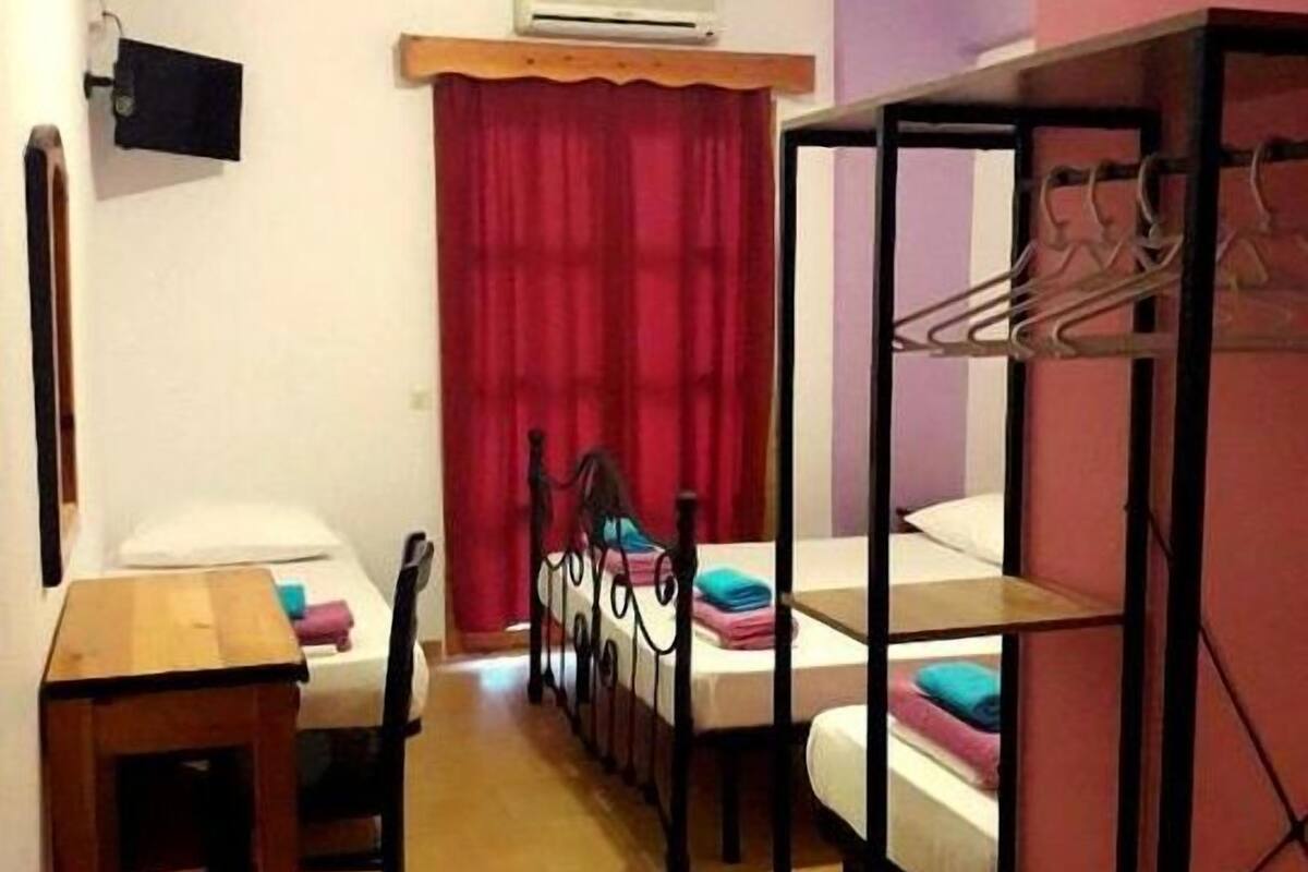A compact room with a bed, a desk, and a chair. There is a red curtain, a wall-mounted air conditioner, and a clothes rack.