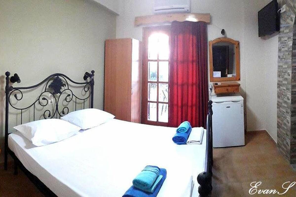A bedroom with a bed, a wooden nightstand, a small desk, a mirror, and a window with red curtains.
