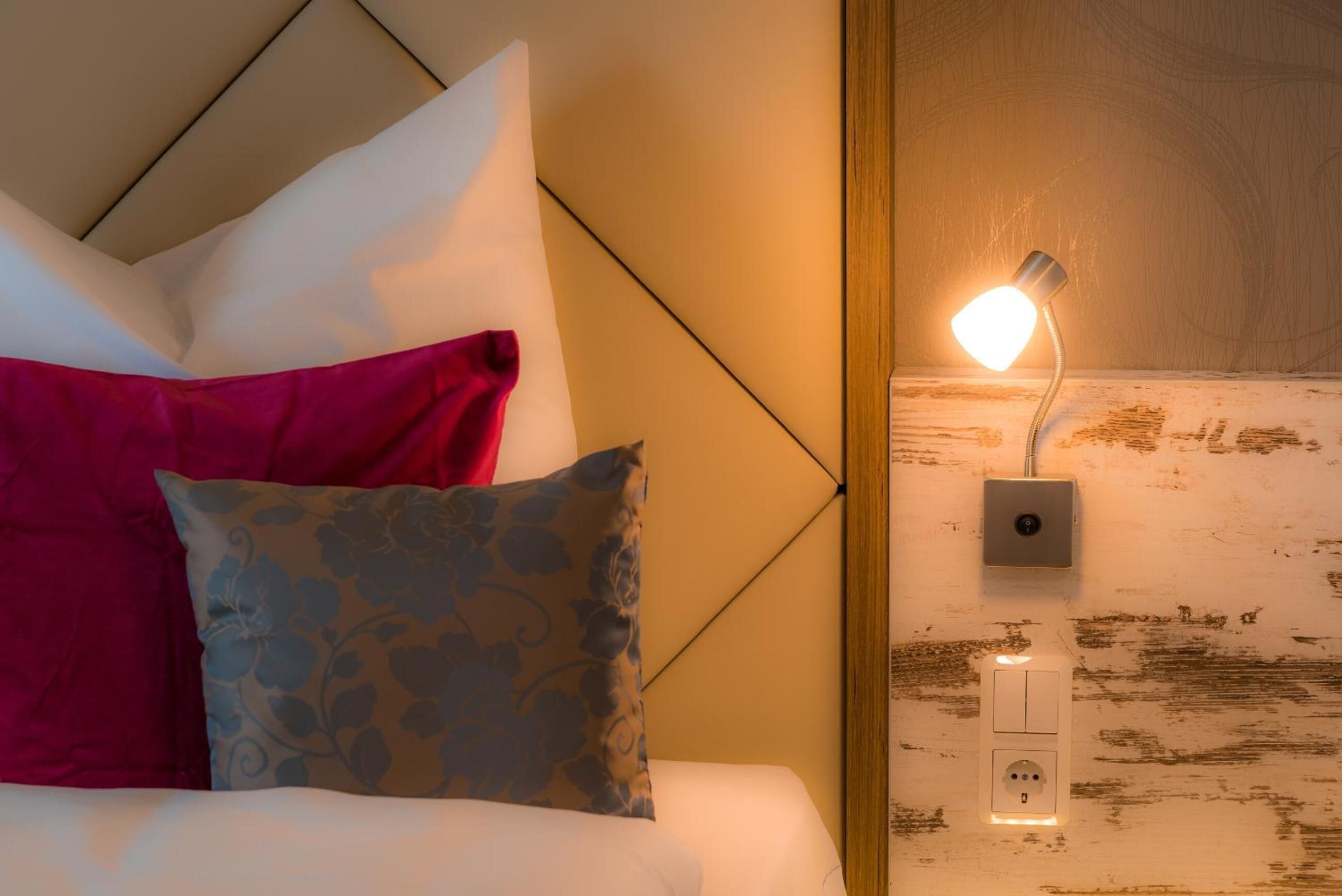 En pænt redt seng med en hvid og lyserød pude, en grå pud med et mønster, og en vægmonteret lampe med en kontakt.