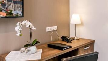 A hotel room with a wooden desk, a lamp, a phone, a small potted orchid, and a framed cityscape picture on the wall.