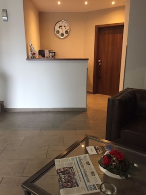 A reception area with a clock, a reception desk, a leather couch, and a glass coffee table with a newspaper and flowers. - Hotel Allegro (Halberstadt)