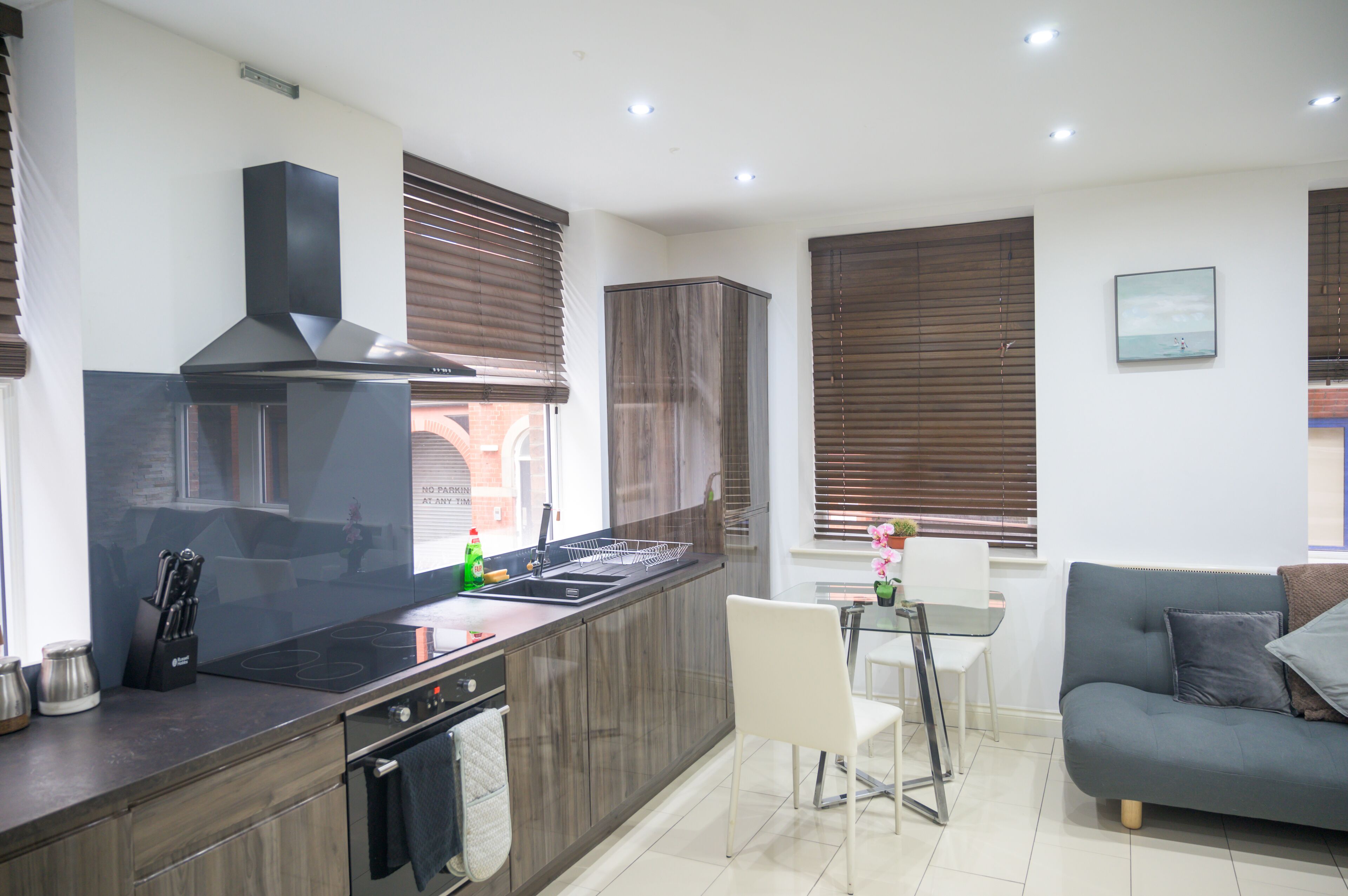A modern kitchen with a central island, built-in oven, and stainless steel appliances. There is a dining area with a white table and chairs, and a grey sofa in the living room.