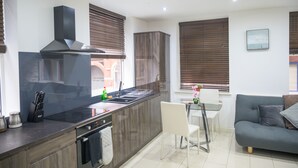Una cocina moderna con una isla central, horno empotrado y electrodomésticos de acero inoxidable. Hay un comedor con una mesa y sillas blancas, y un sofá gris en la sala de estar.