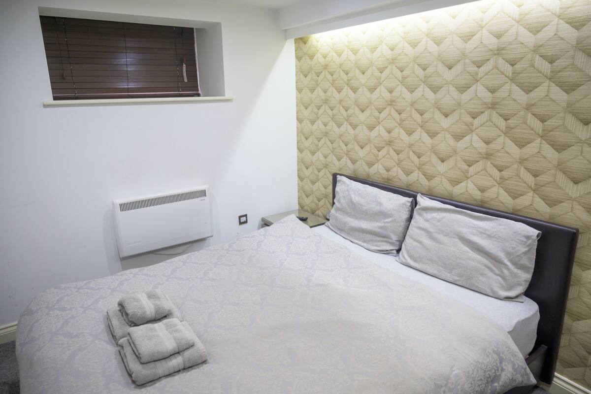 A neatly made bed with a quilted headboard, a window with blinds, and a wall-mounted air conditioner.