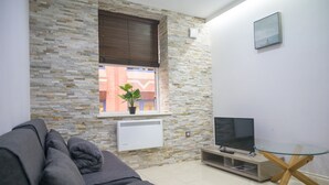 Una sala de estar con pared de piedra, una ventana con persianas, un sofá gris, una mesa de centro de vidrio y un televisor de pantalla plana.