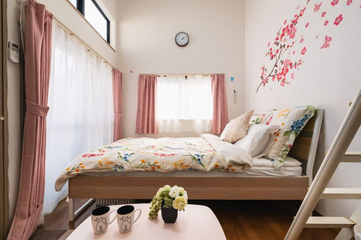 Ein Schlafzimmer mit einem Bett, einem Nachttisch mit Blumenvase, einer Wanduhr und einem Fenster mit Vorhängen.