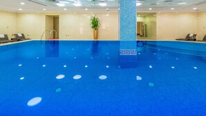 A large indoor swimming pool with clear blue water, lounge chairs, and a potted plant. - Al Diyafa Furnished Suites (Doha)