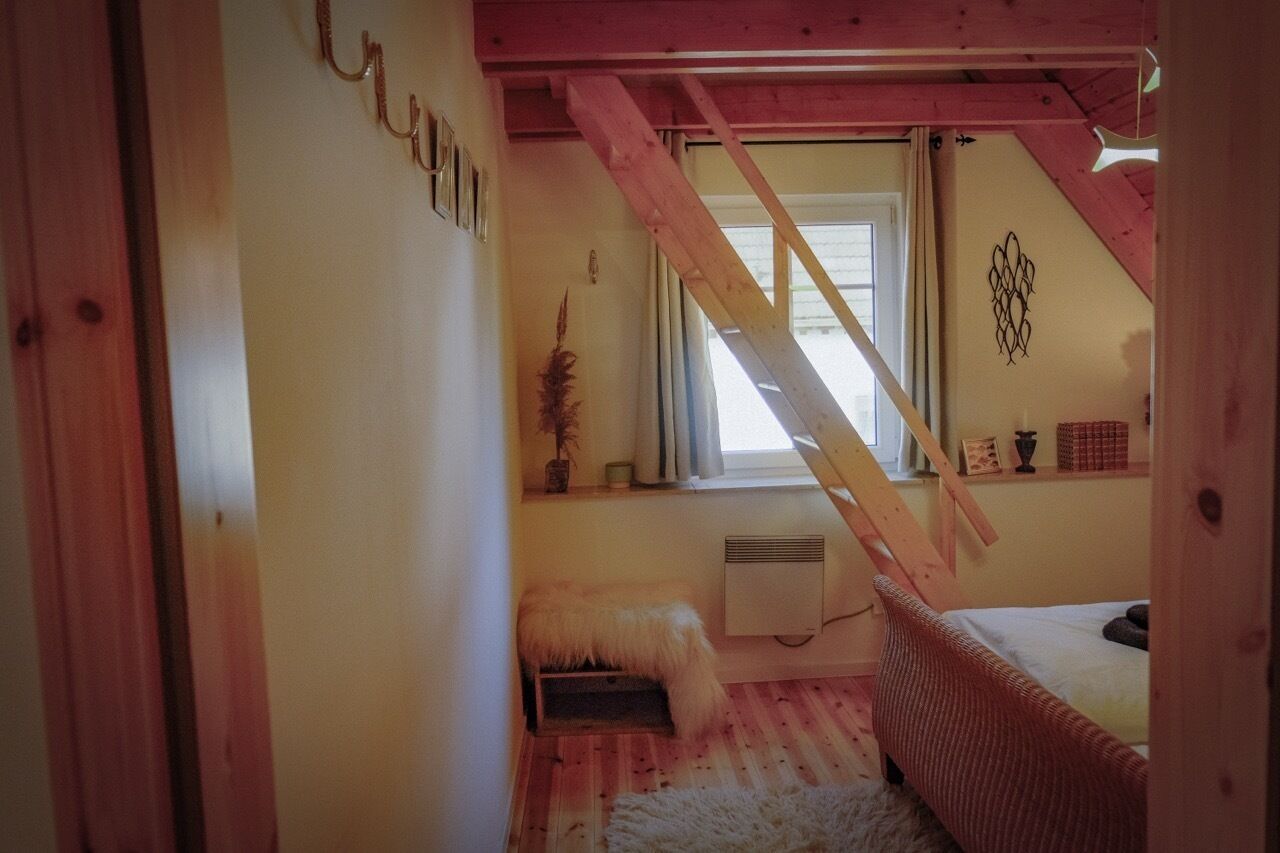 Un dormitorio acogedor en el ático, con techo inclinado, una escalera de madera, una cama, un pequeño banco y un radiador.