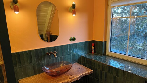 Une salle de bain avec un meuble-lavabo en bois, un miroir rond et une fenêtre donnant sur les arbres.