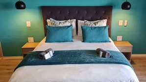 A neatly made bed with a teal bedspread, two blue pillows, and a dark headboard. There are two bedside tables with lamps, and a wooden nightstand with a lamp.