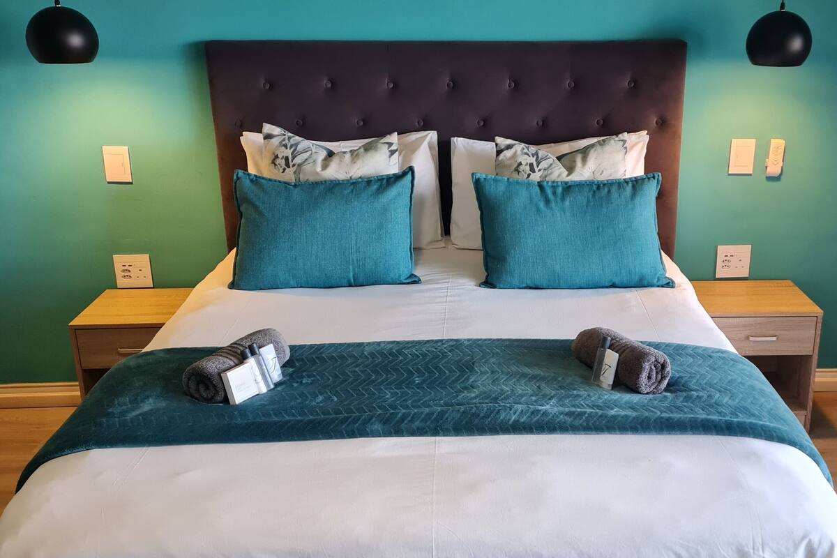 A neatly made bed with a teal bedspread, two blue pillows, and a dark headboard. There are two bedside tables with lamps, and a wooden nightstand with a lamp.