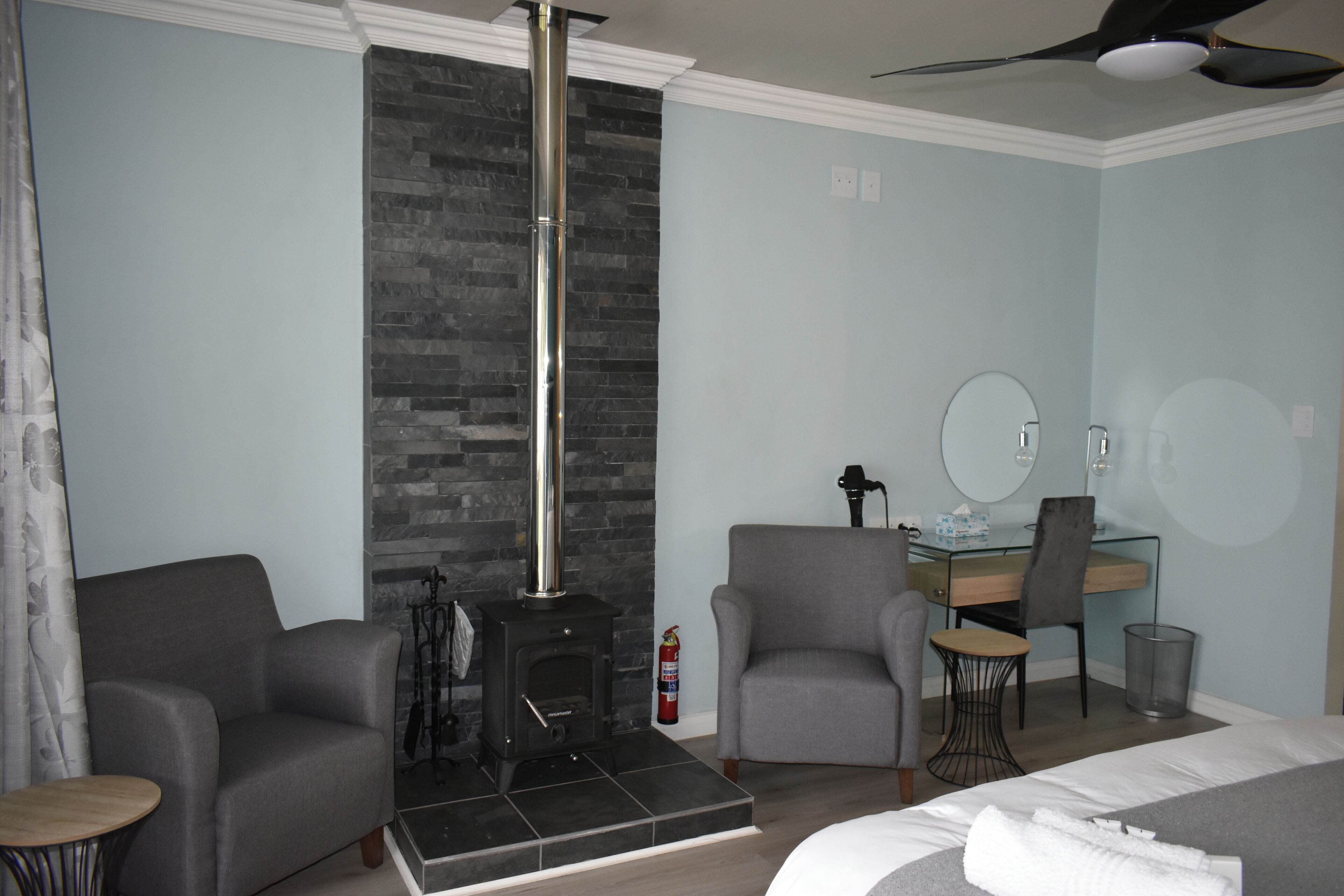 Una habitación de hotel con chimenea, dos sillones, un escritorio con silla, una cama y un ventilador de techo.