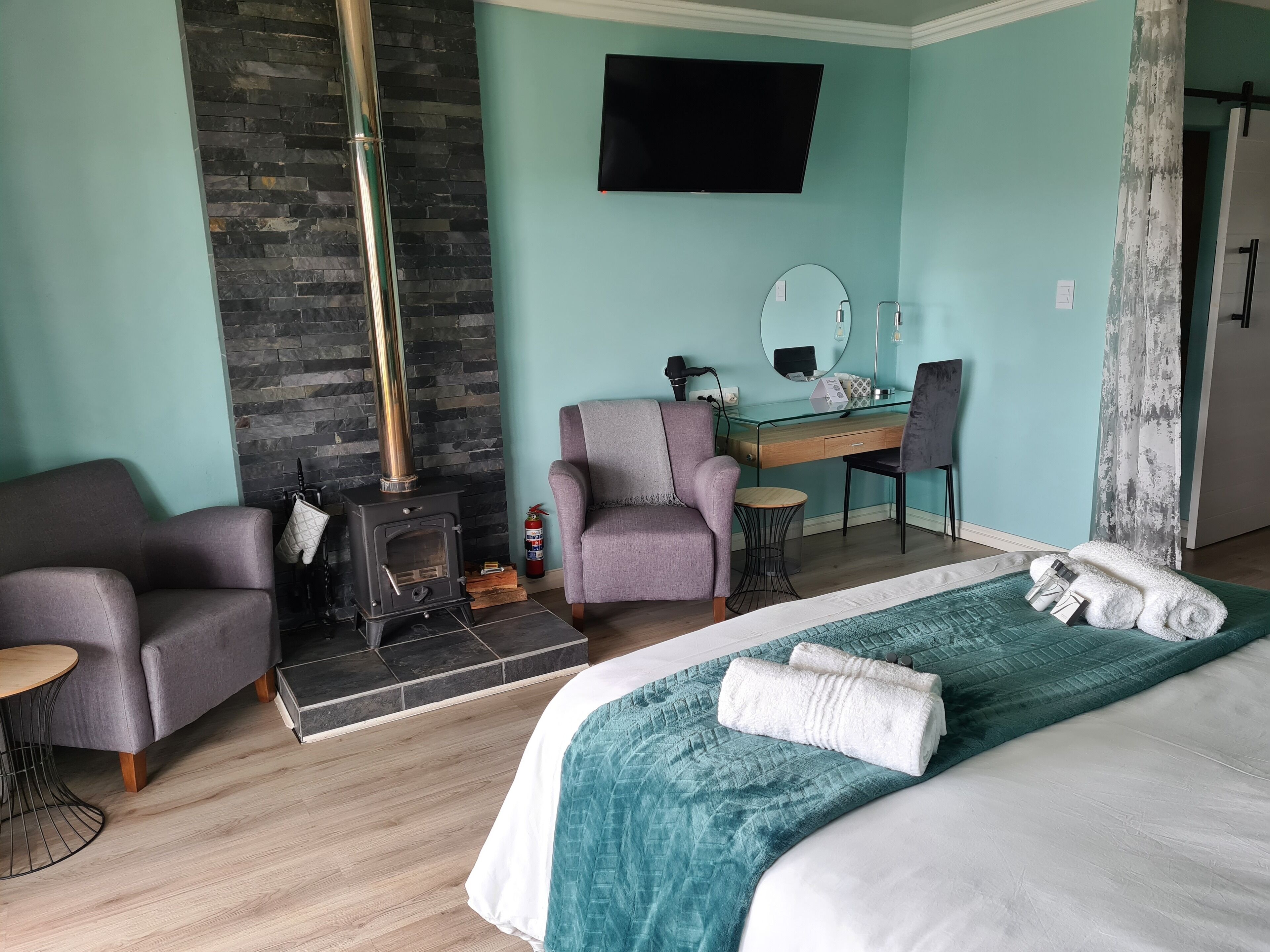 A modern hotel room with a stone fireplace, a desk with a mirror, a comfortable armchair, and a neatly made bed.