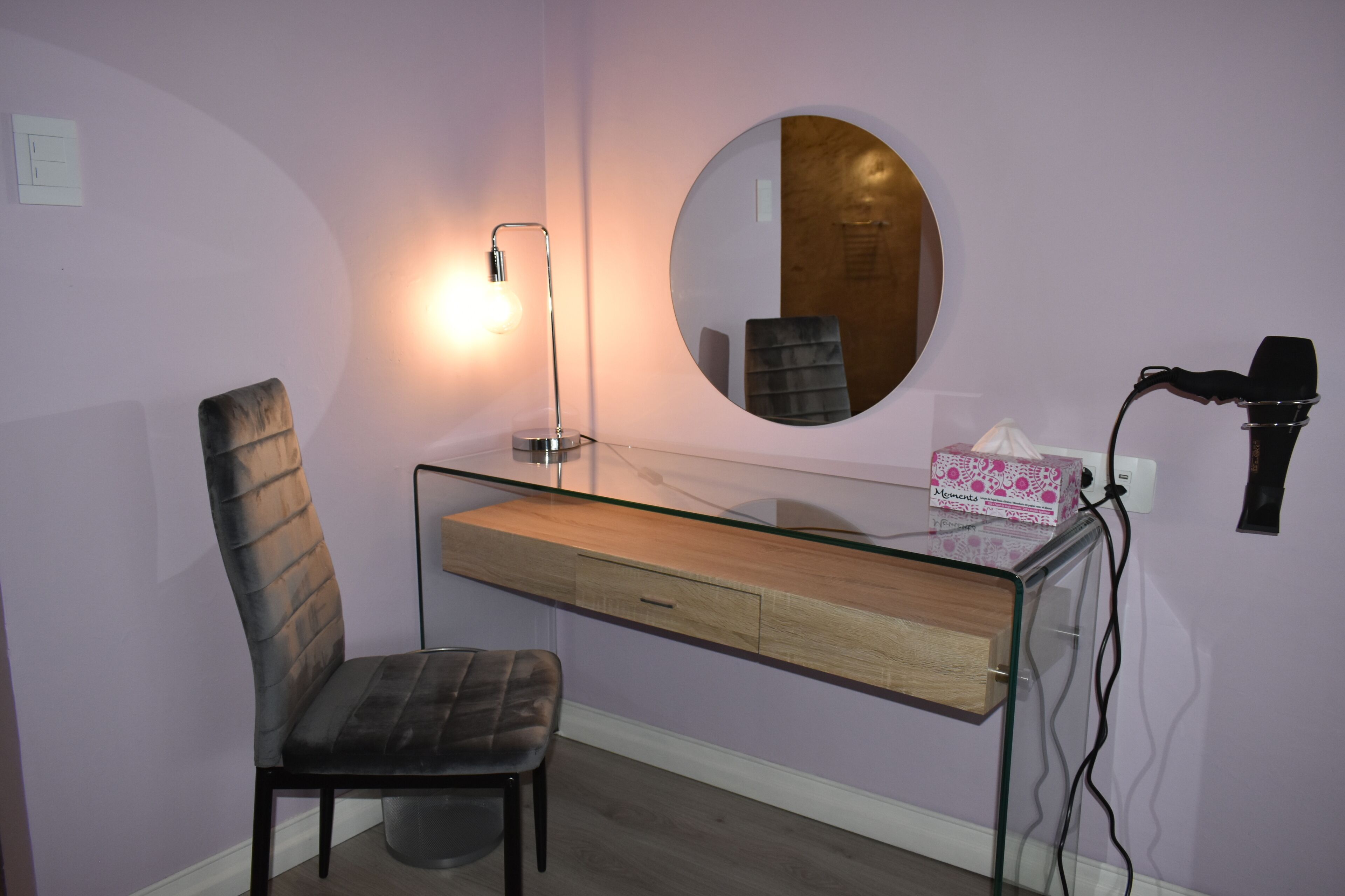 A room with a glass vanity, a chair, a round mirror, a hairdryer, and a lamp.