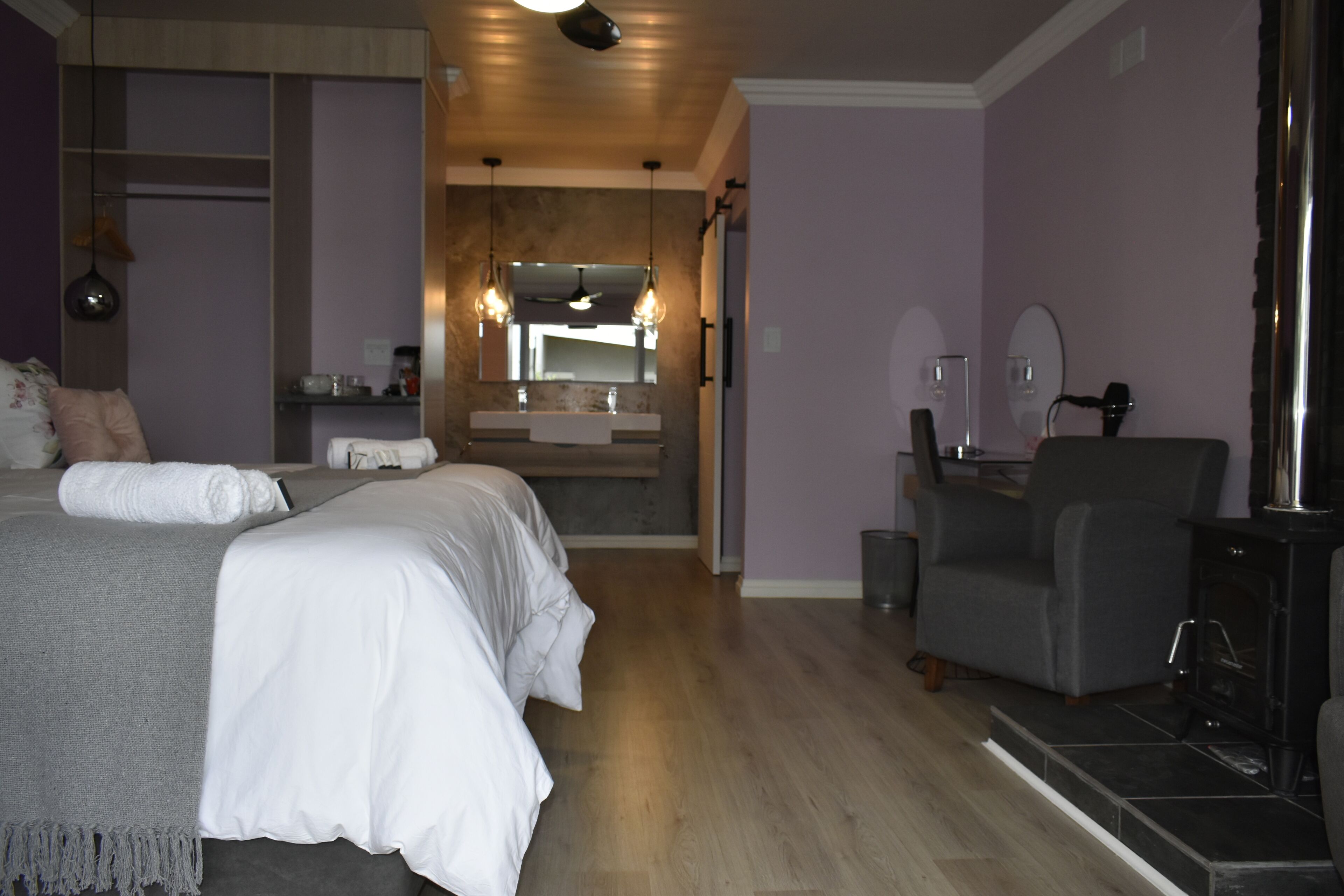 Habitación de hotel moderna con una cama grande, un sillón gris, una chimenea y un baño a lo lejos.