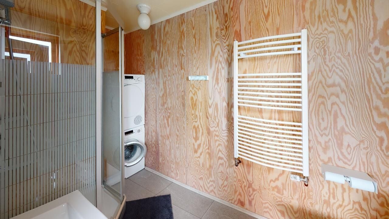 A bathroom with a wooden wall, a white radiator, a washing machine, and a shower area.