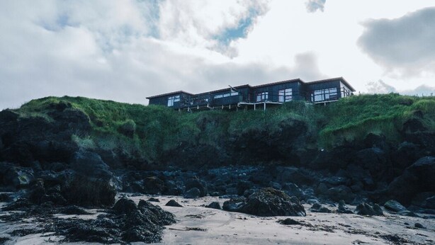 A modern building with large windows, situated on a grassy hill with a rocky foreground.