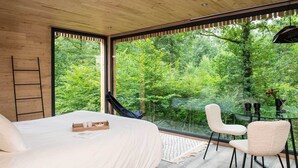 Une chambre à coucher avec un lit, une échelle, une table en verre avec des chaises, et une vue sur une forêt à travers de grandes fenêtres.