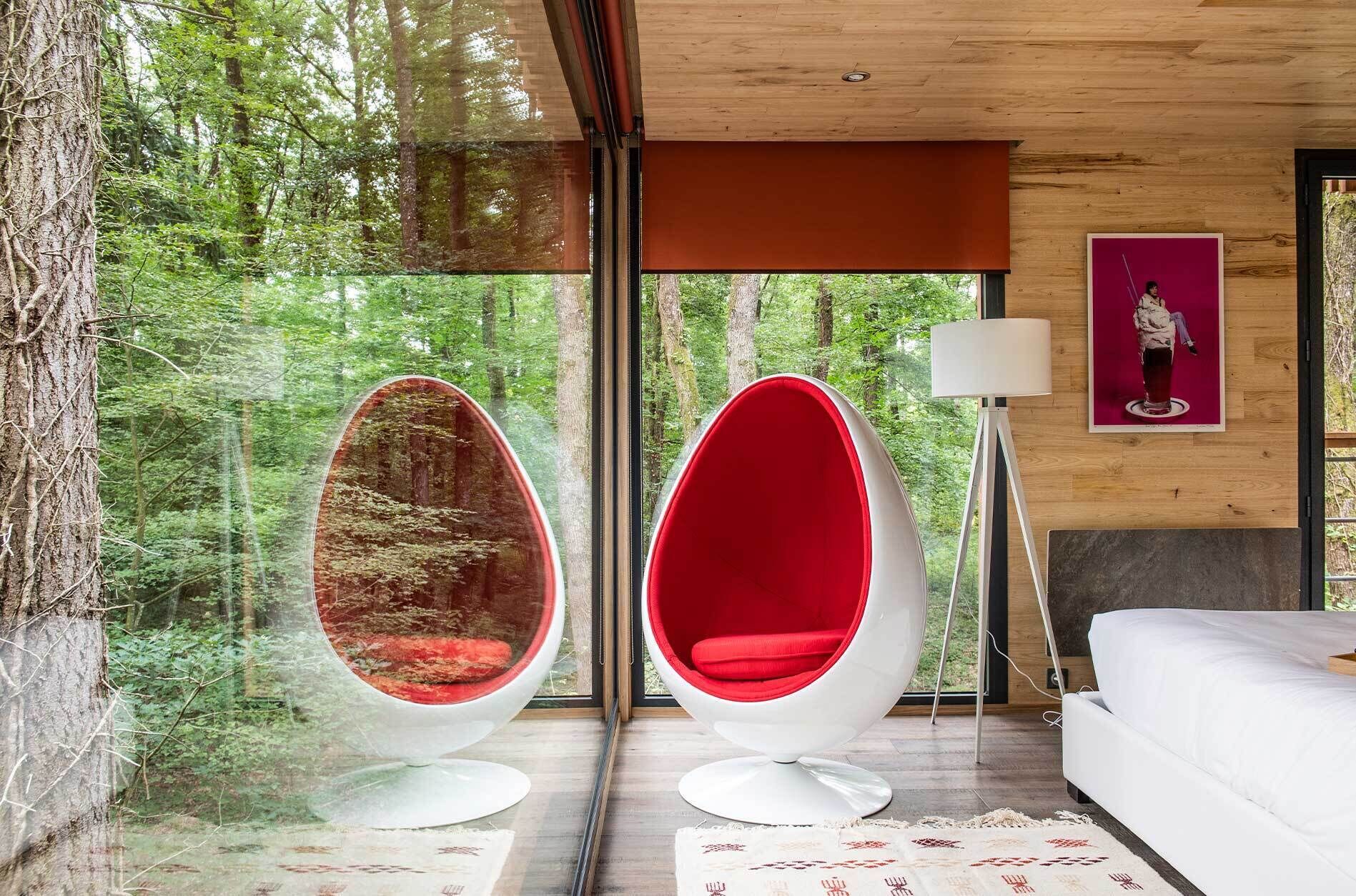 Un dormitorio moderno con un ventanal grande, dos sillones con forma de huevo, una cama y una lámpara de pie.