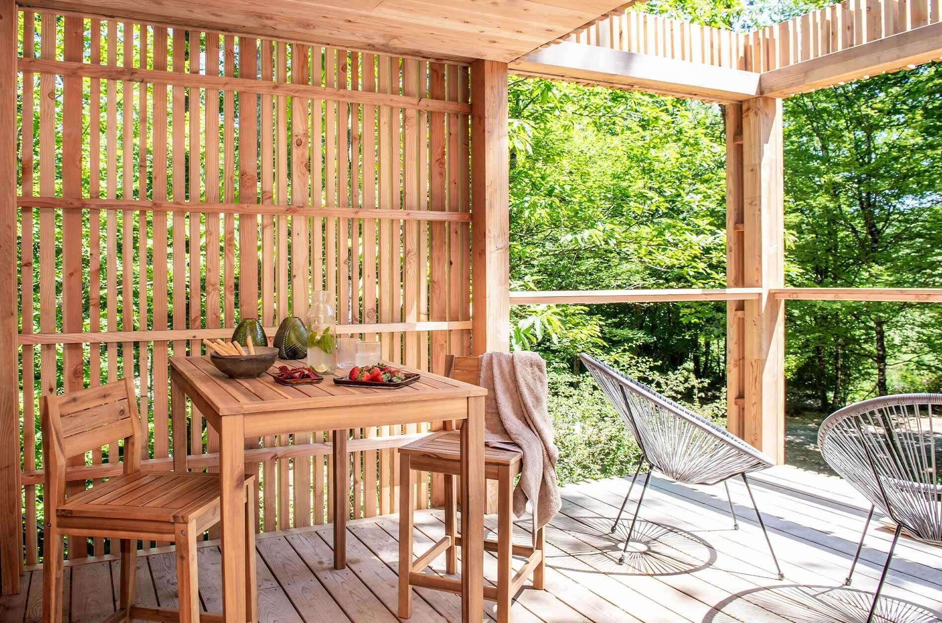 Una terraza de madera al aire libre con una mesa y sillas, rodeada por un cerco de madera y grandes ventanales que dan a vistas de vegetación.