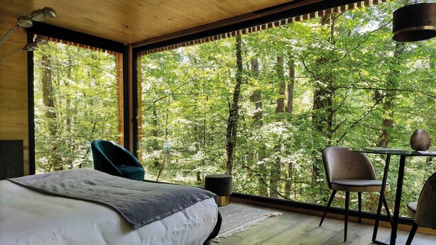 Une chambre avec une grande fenêtre, un lit, une chaise, une petite table et une lampe.