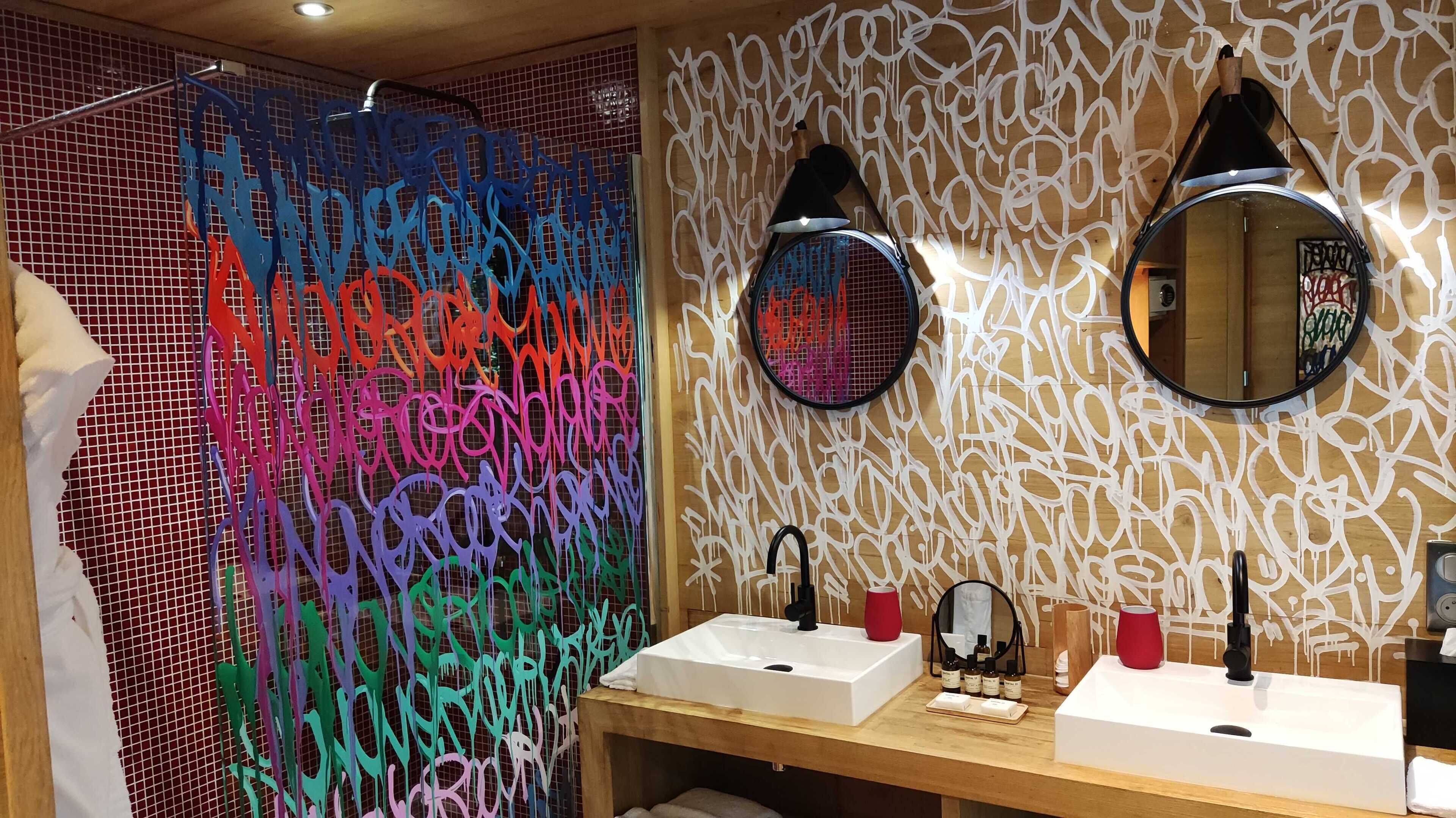 A modern bathroom with a shower area featuring colorful, abstract wall art. There are two sinks with mirrors above them, and a wooden vanity with toiletries.