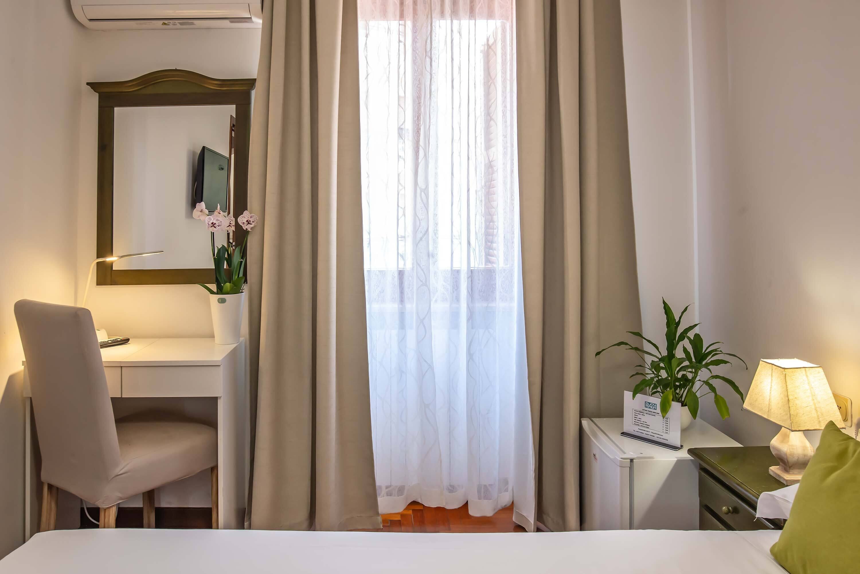 A hotel room with a bed, a desk, a chair, a lamp, and a window with curtains.
