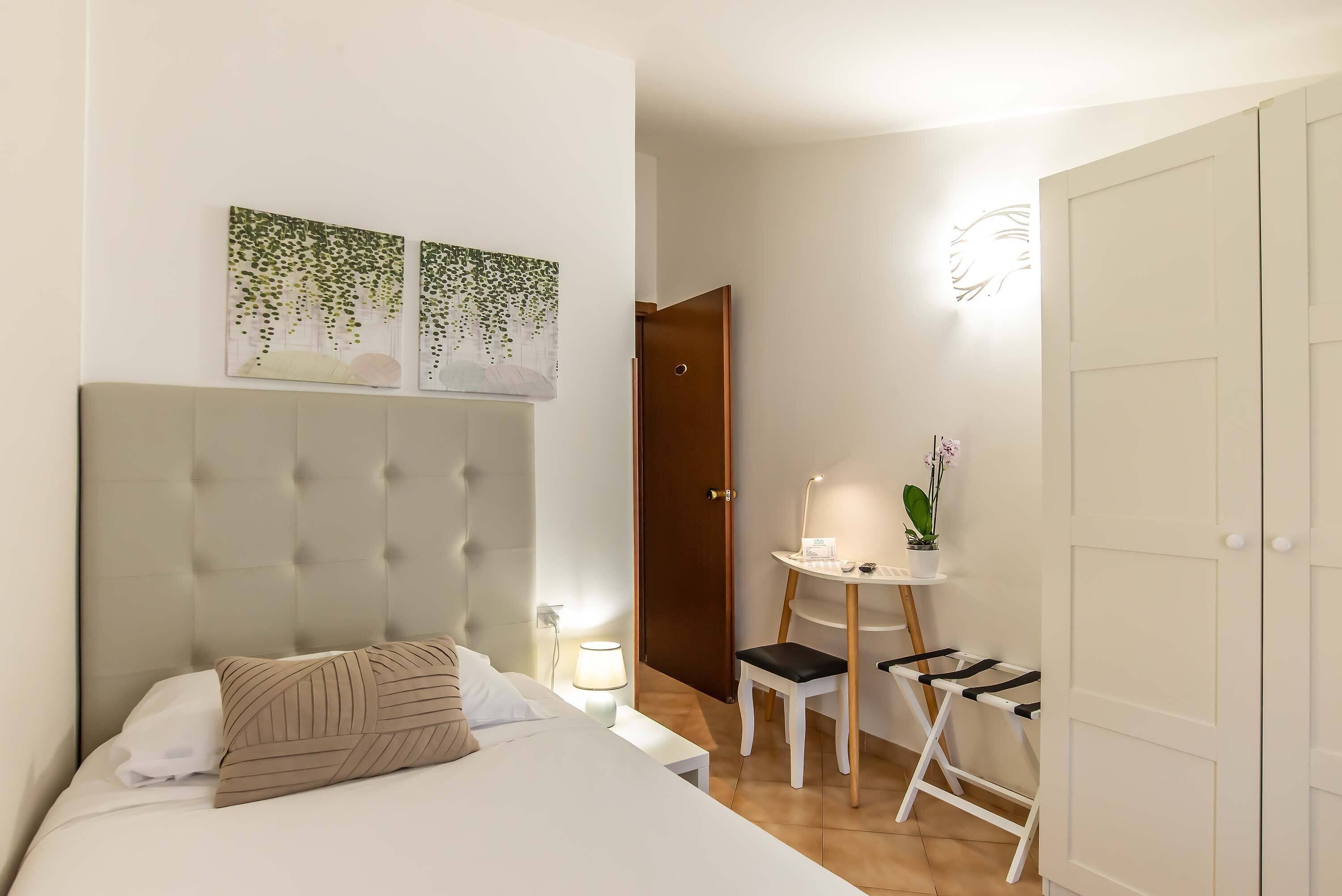 A hotel room with a tufted headboard bed, a small table with a lamp, a chair, and a potted plant.