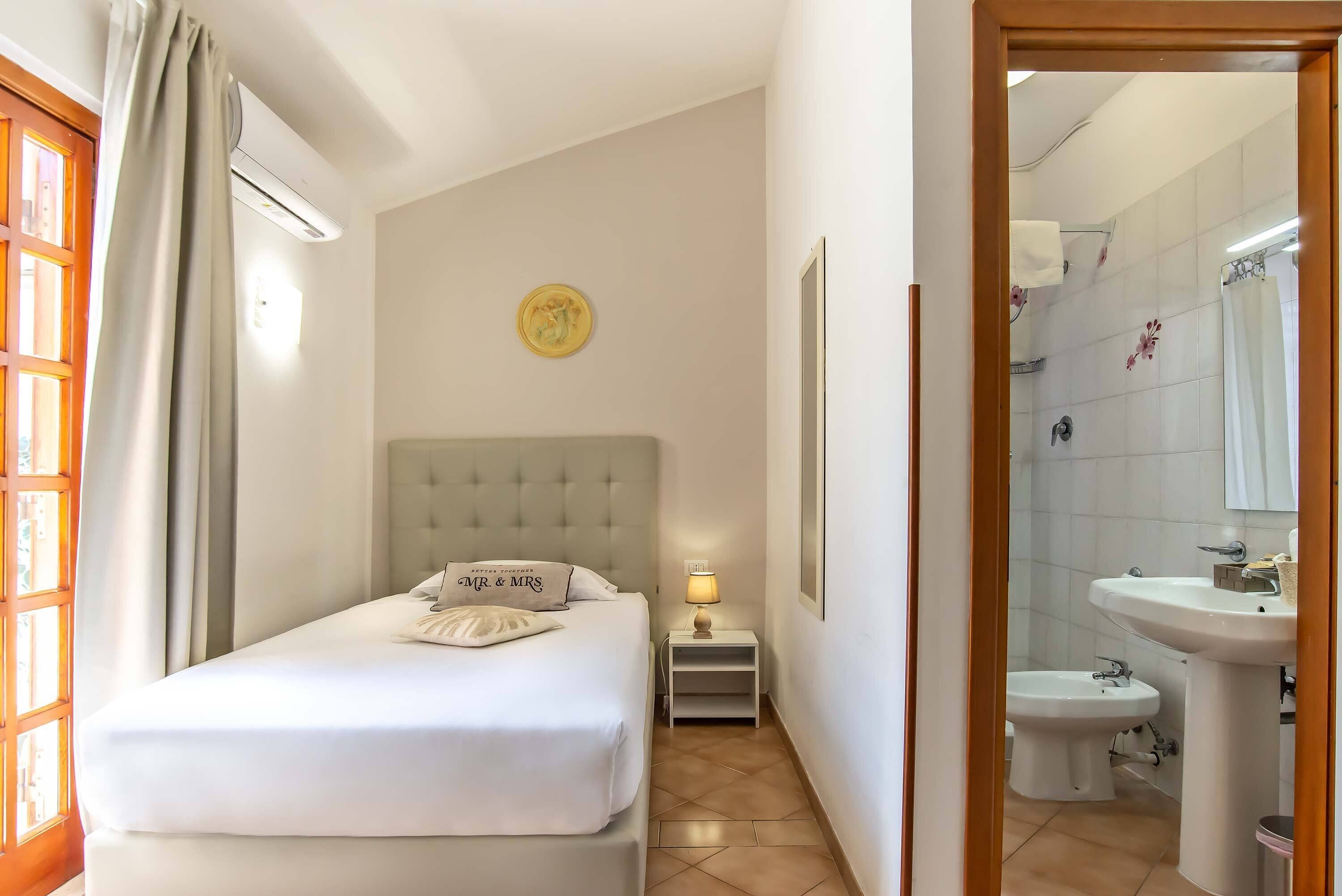 A hotel room with a bed, a bedside table with a lamp, a small shelf, a wall clock, and a bathroom with a sink and toilet.