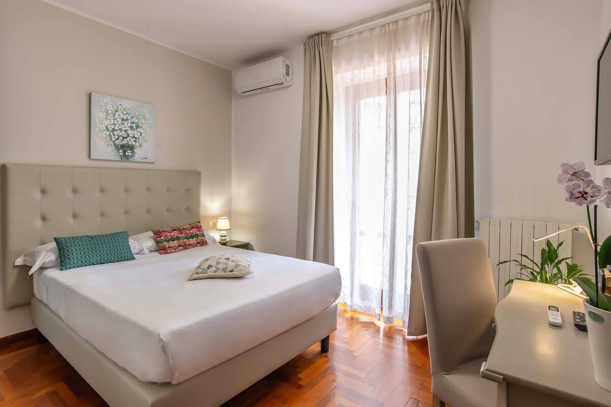 A hotel room with a large bed, a bedside table with a lamp, a chair, a potted plant, and a window with curtains.