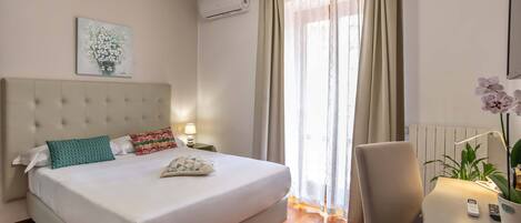 A hotel room with a large bed, a bedside table with a lamp, a chair, a potted plant, and a window with curtains.