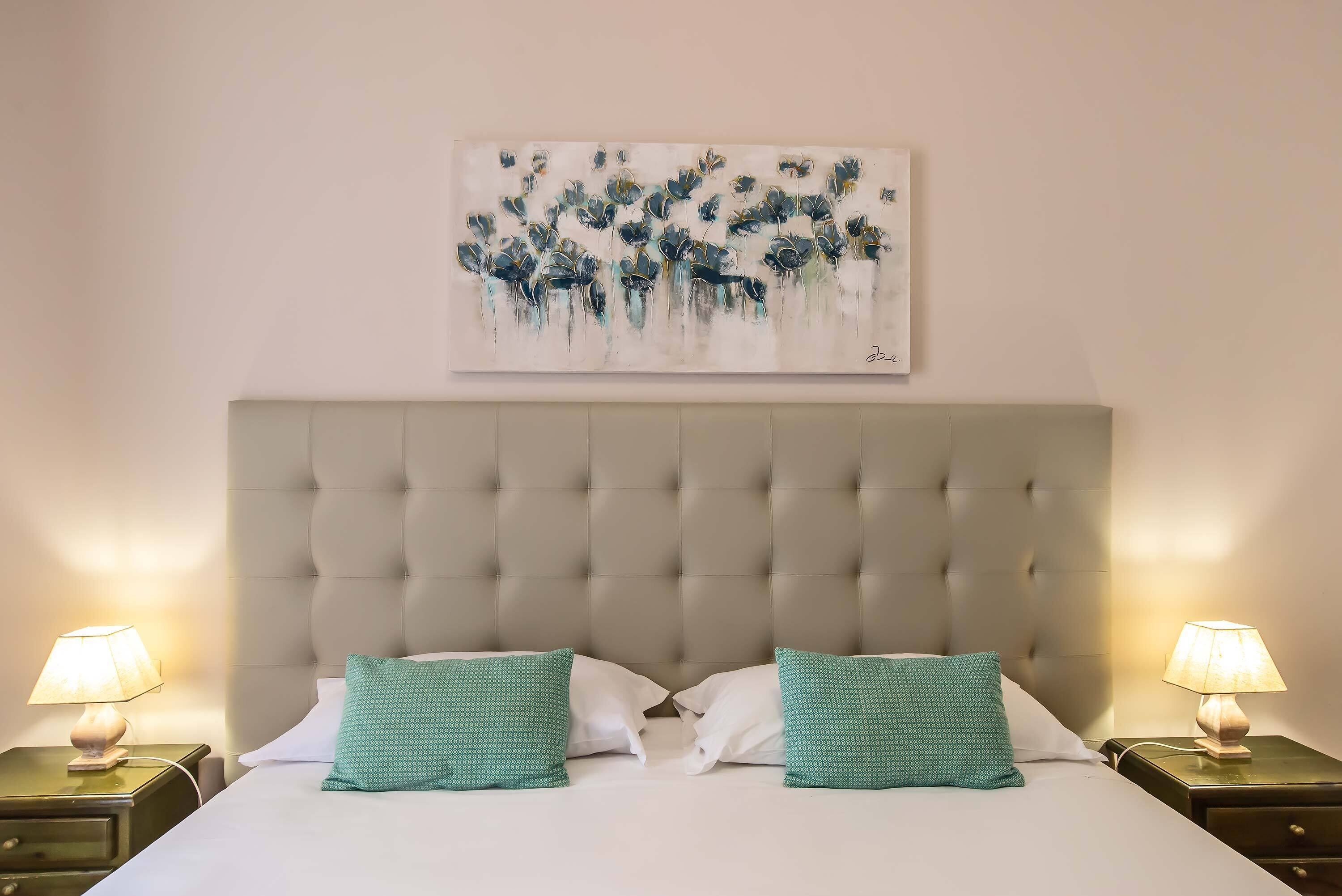 Hotelkamer met een gestuufd hoofdeinde, twee groene kussens met een patroon, een wit sprei en een schilderij boven het bed.