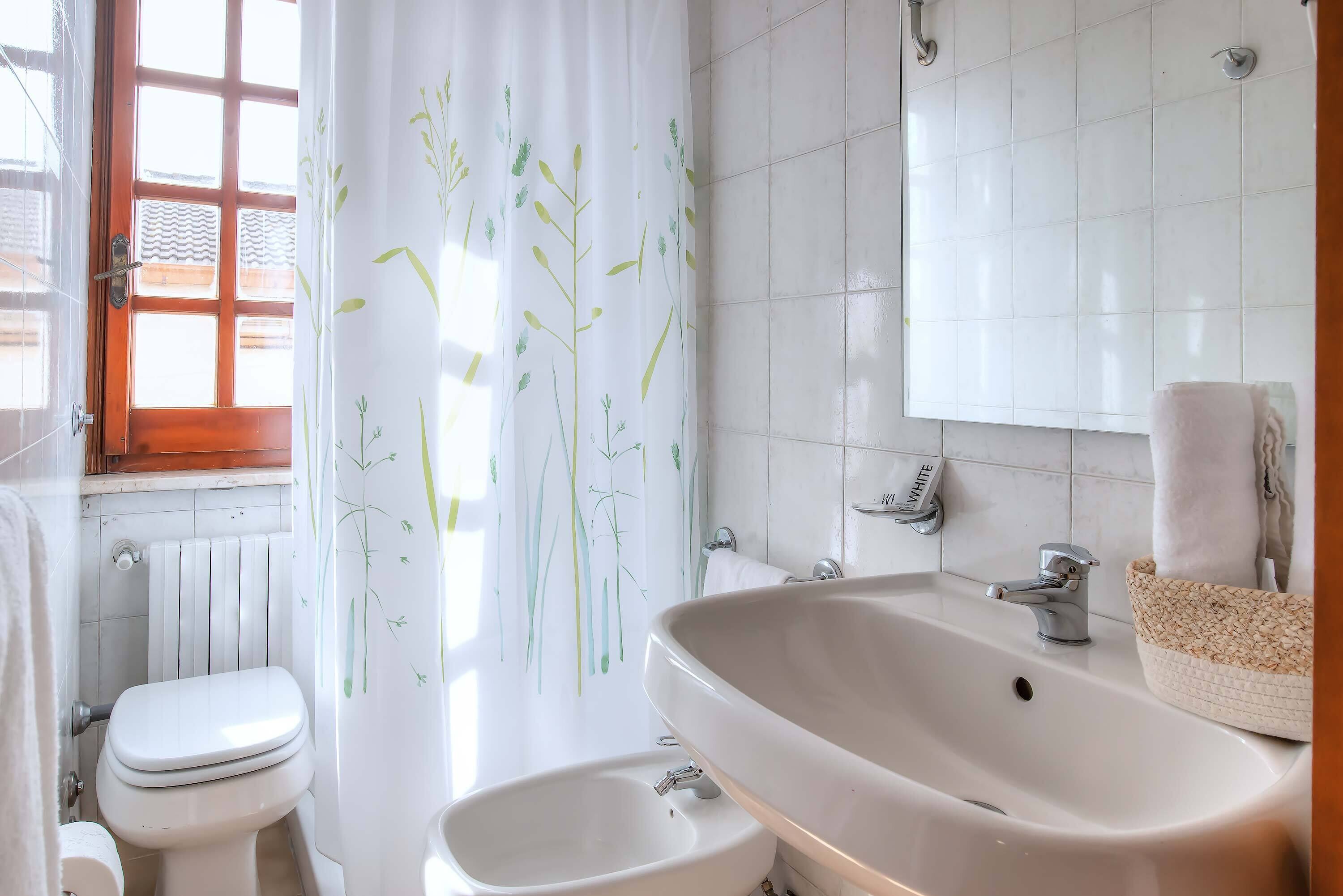 A bathroom with a toilet, bidet, sink, and a window with a wooden frame and white curtains with a plant design.