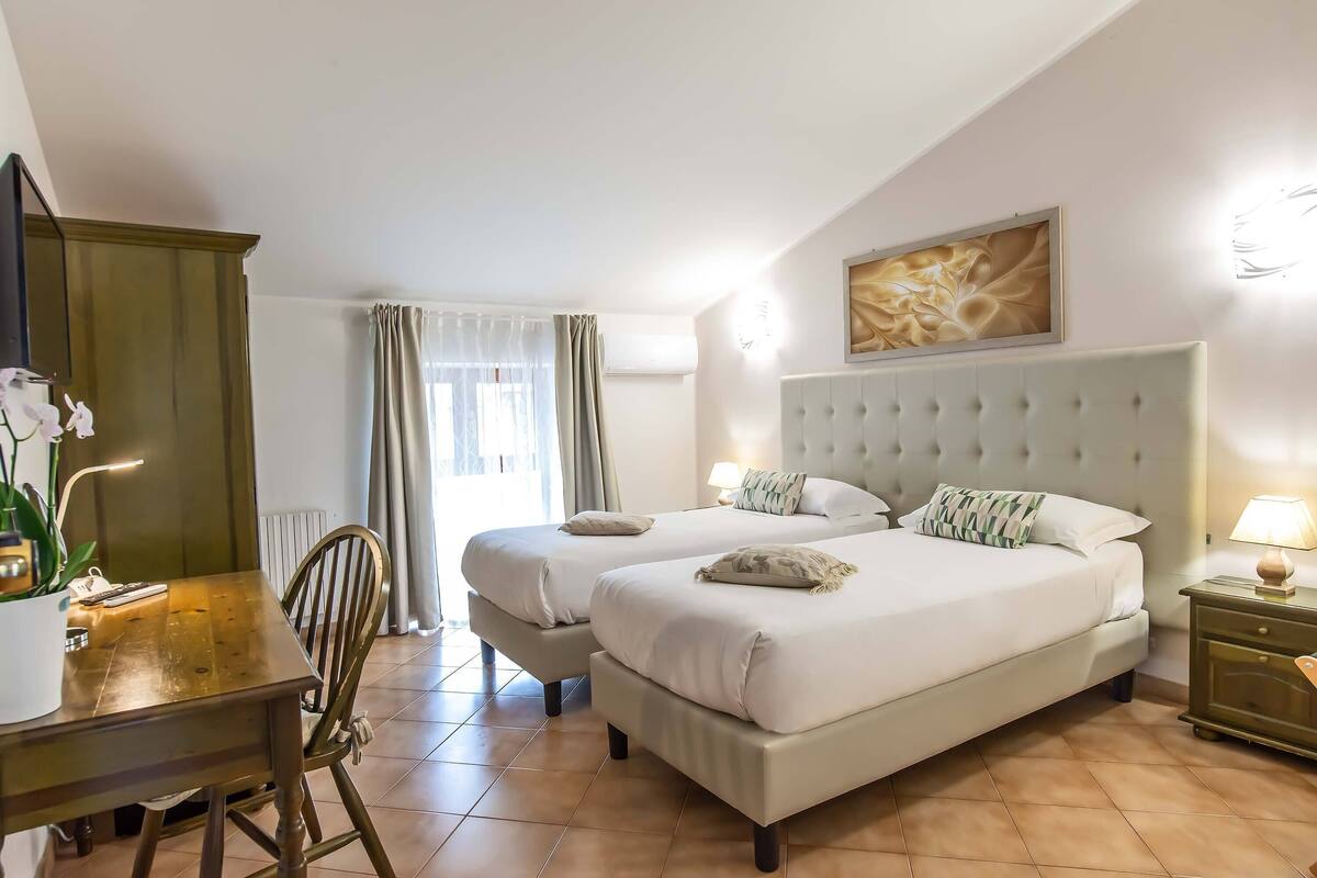 A hotel room with two beds, a wooden desk, a chair, a potted plant, and a window with curtains.