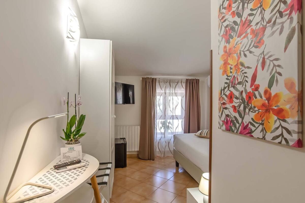 A hotel room with a bed, a desk with a computer and a plant, a TV mounted on the wall, and a window with curtains.
