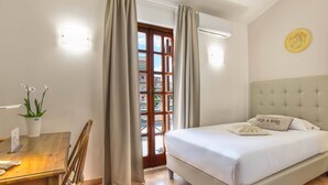 A bedroom with a bed, a desk with a chair, a potted plant, and a window with curtains. - Plaza Rooms Ciampino (Ciampino)