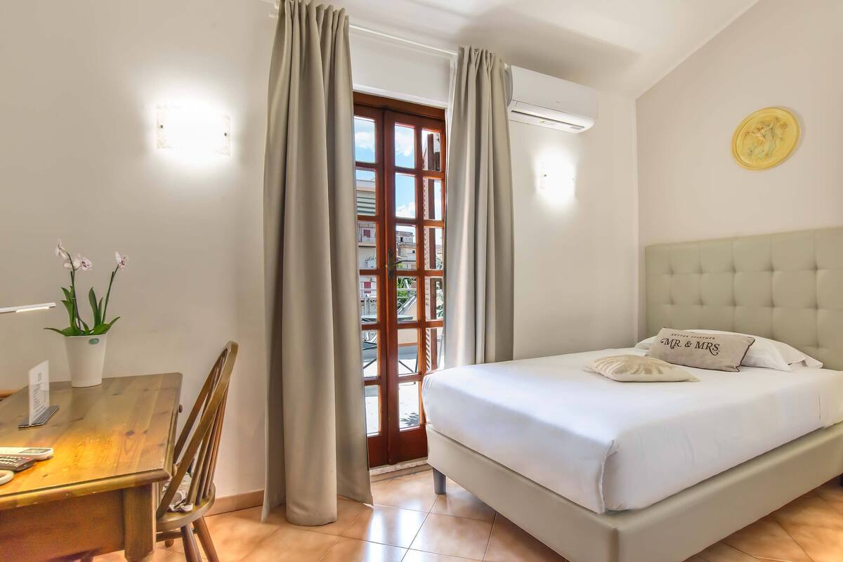 A bedroom with a bed, a desk with a chair, a potted plant, and a window with curtains.