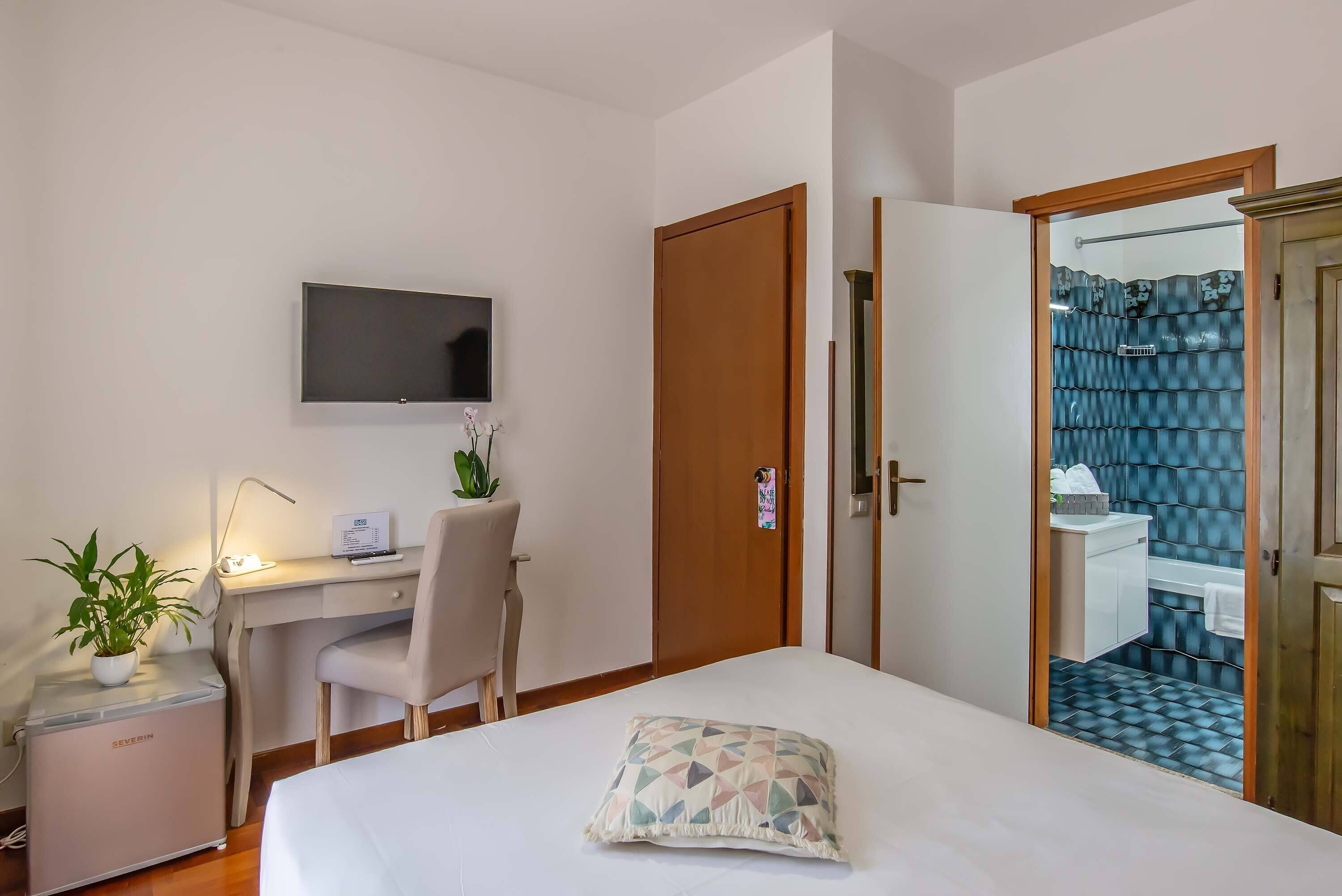 A hotel room with a bed, a desk with a chair, a television, a small refrigerator, and a bathroom visible through an open door.