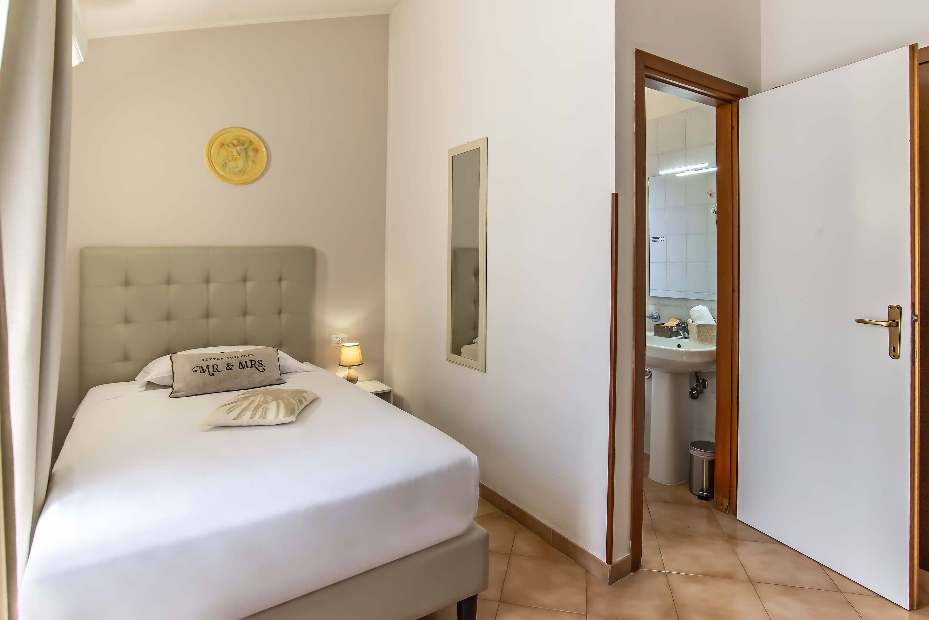 A bedroom with a bed, a bedside lamp, a mirror, and a door leading to a bathroom.