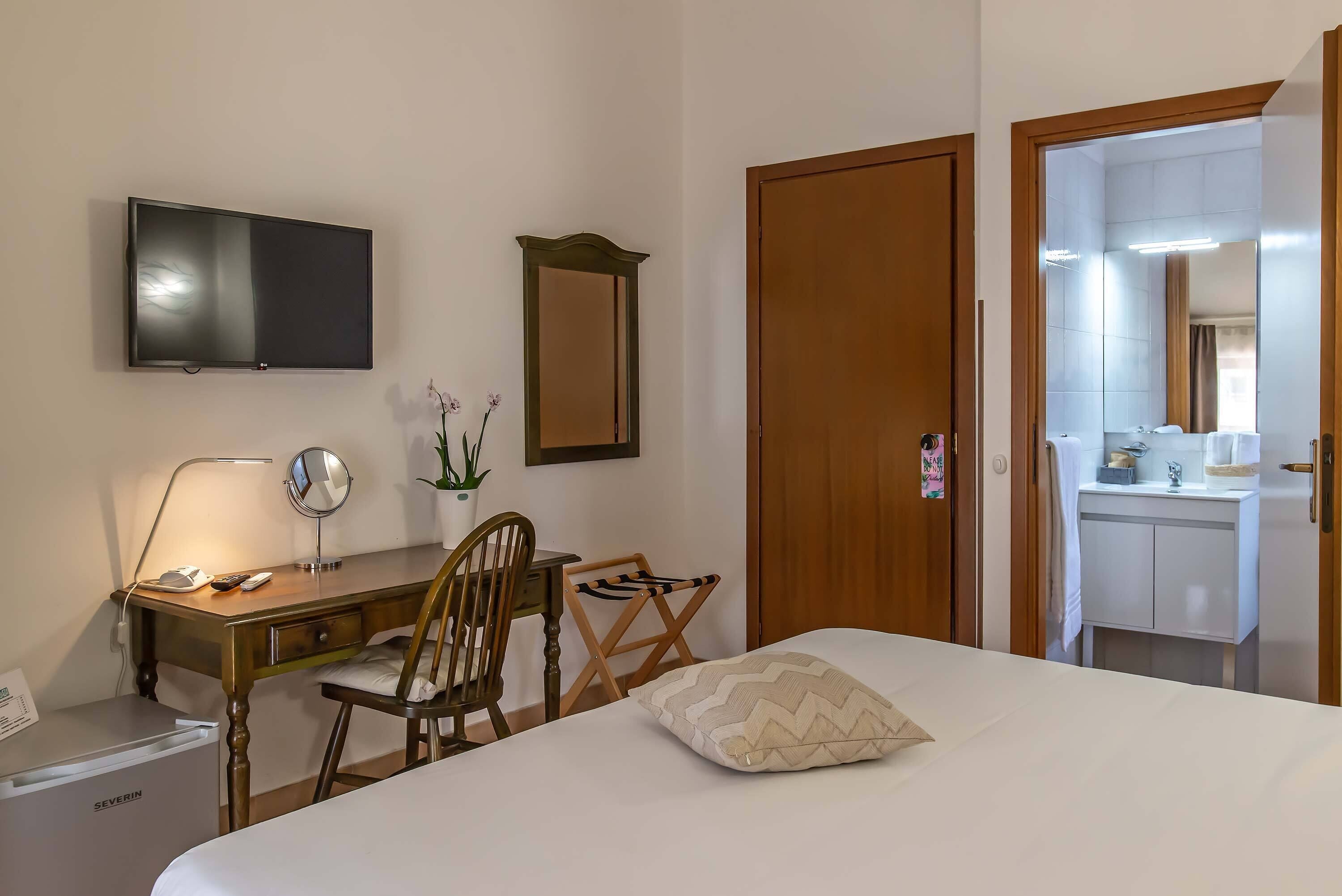 A hotel room with a bed, a desk with a chair, a TV, a mirror, a small fridge, and a bathroom visible through an open door.