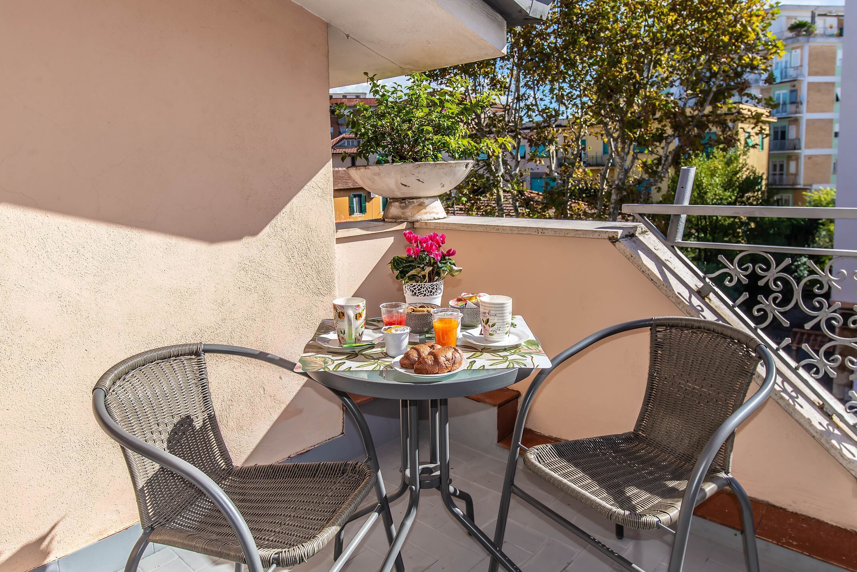 A balcony with a table set for breakfast, two wicker chairs, a potted plant, and a railing.