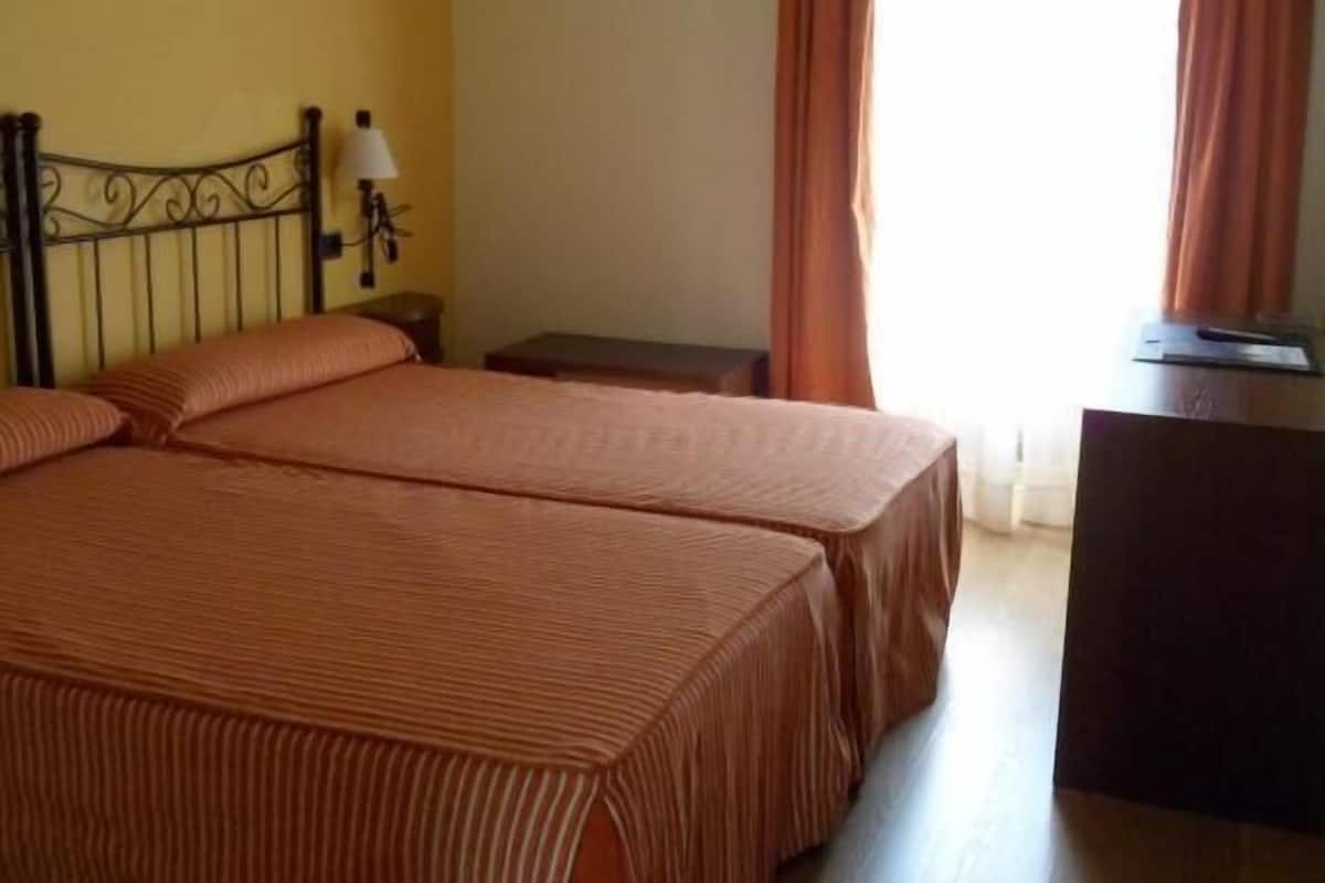 A double bed with striped bedding, a nightstand with a lamp, and a TV on a wooden dresser in a room with yellow walls and orange curtains.