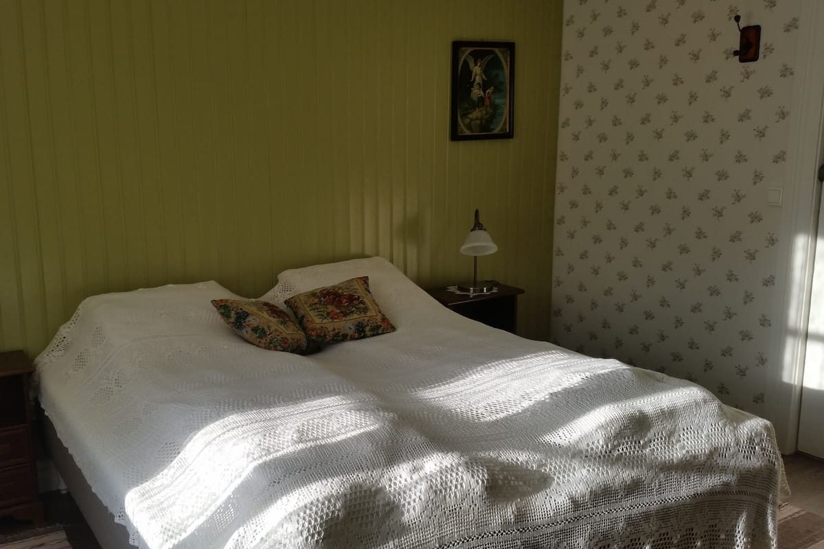 Uma cama bem arrumada com manta branca e almofadas florais, uma mesa de cabeceira com abajur e um quadro na parede.