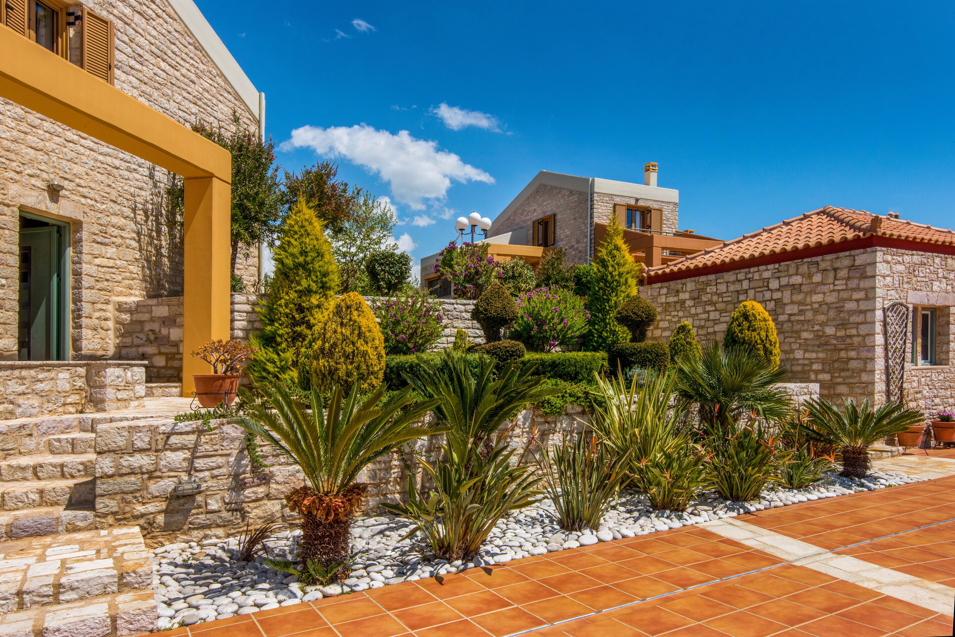 A stone building with a tiled patio, surrounded by various plants and shrubs.