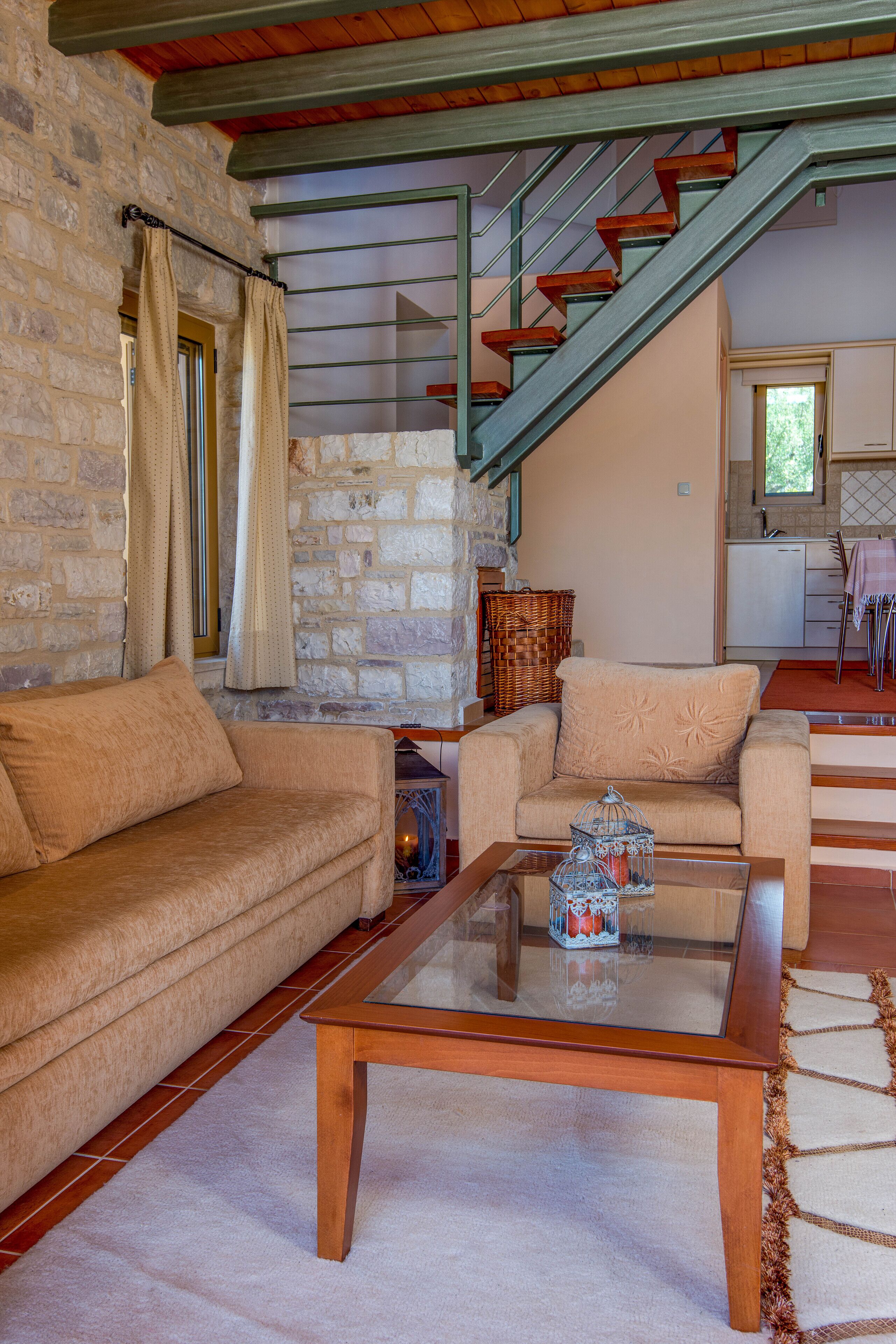 A cozy living room with a stone wall, a wooden staircase, a sofa, a coffee table, and a kitchen in the background.