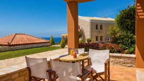 An outdoor patio with a table and chairs, a view of the sea, and a stone building in the background. - Enastron Villas (Trifylia)