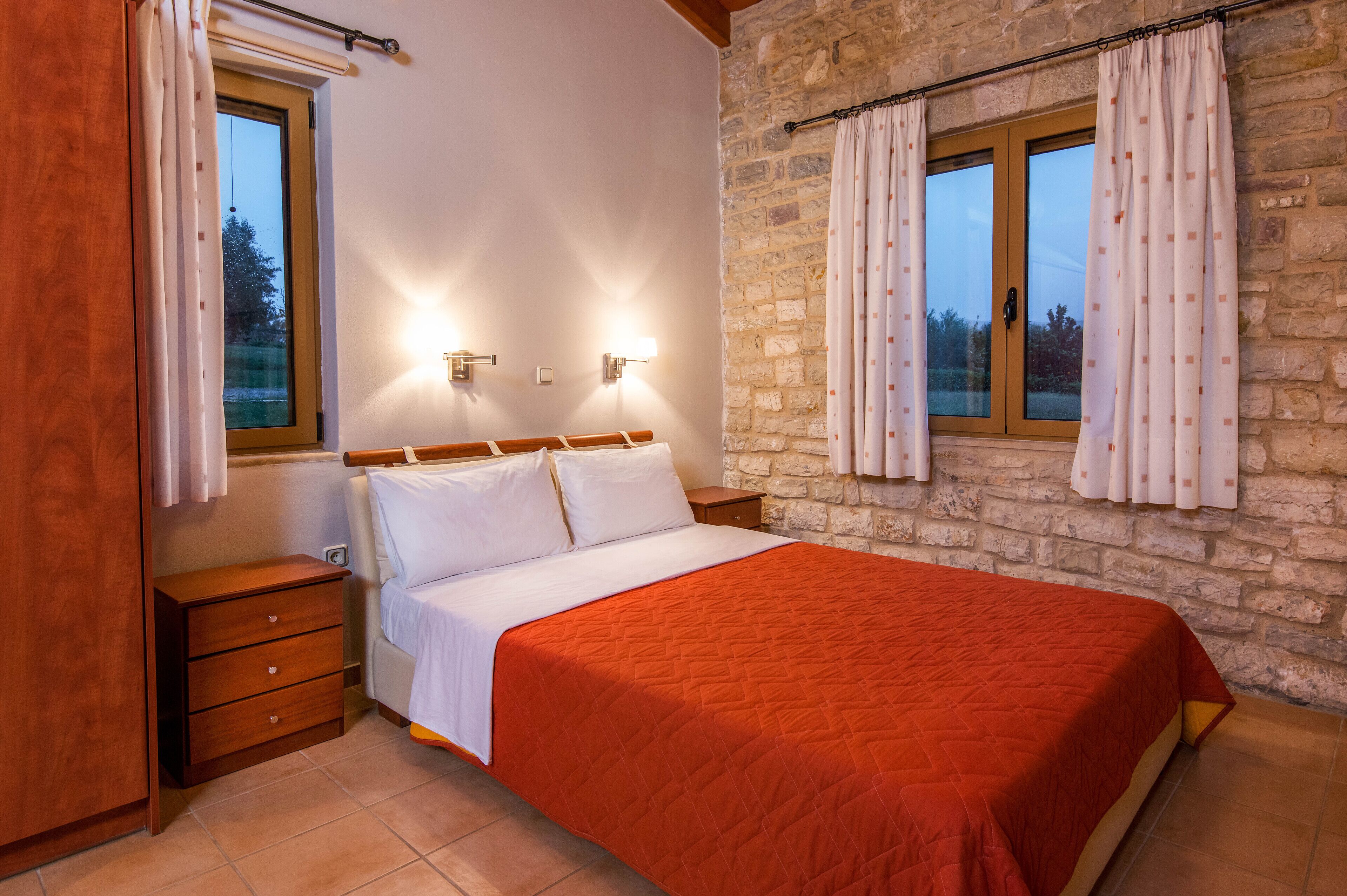 Un dormitorio con cabecera de madera, colcha roja, dos mesitas de noche y una pared de piedra.