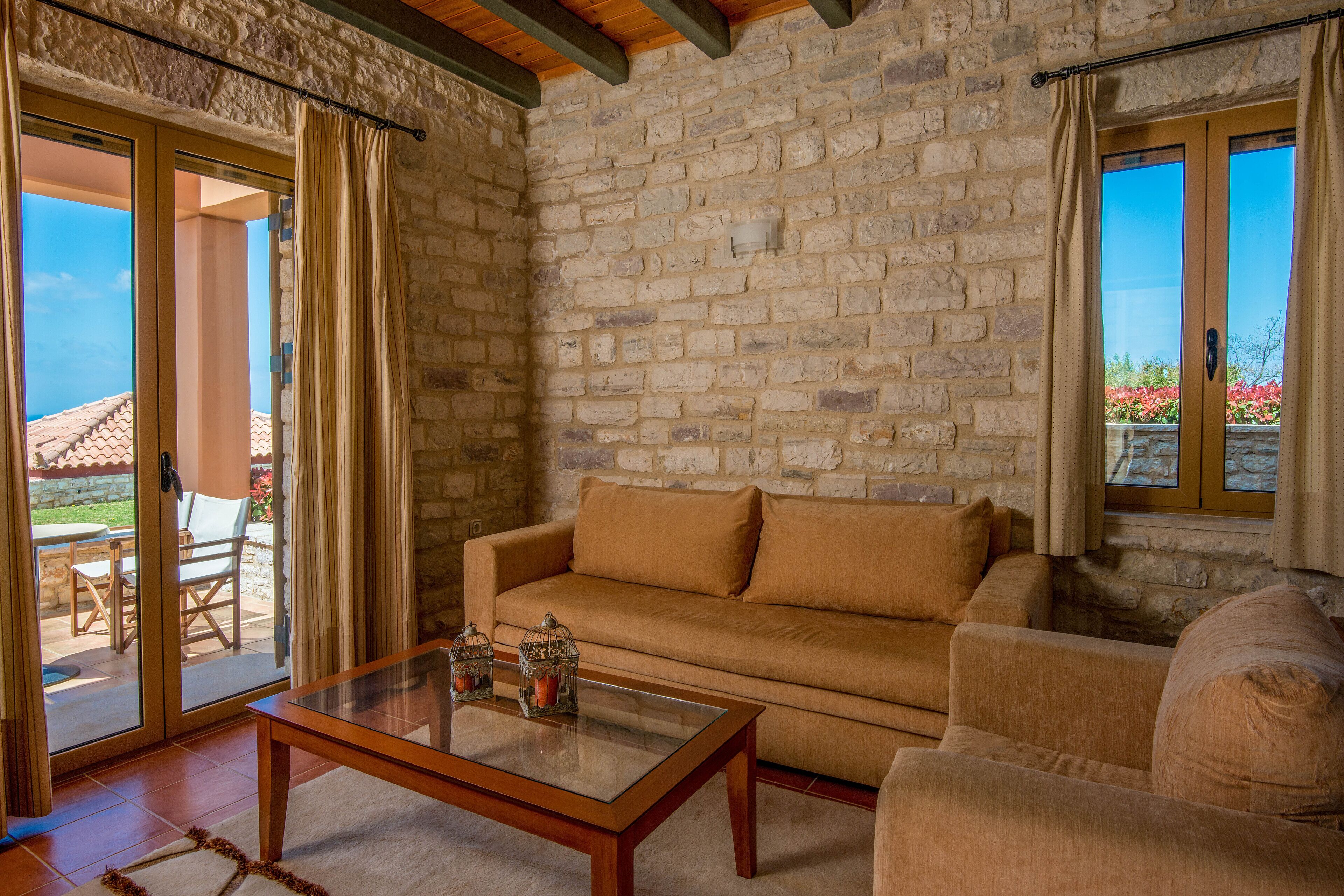 Una habitación con paredes de piedra, un sofá color beige, una mesa de centro de vidrio y un ventanal corredizo que da a un área exterior con un patio y sillas.