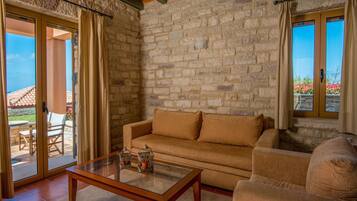 Una habitación con paredes de piedra, un sofá color beige, una mesa de centro de vidrio y un ventanal corredizo que da a un área exterior con un patio y sillas.